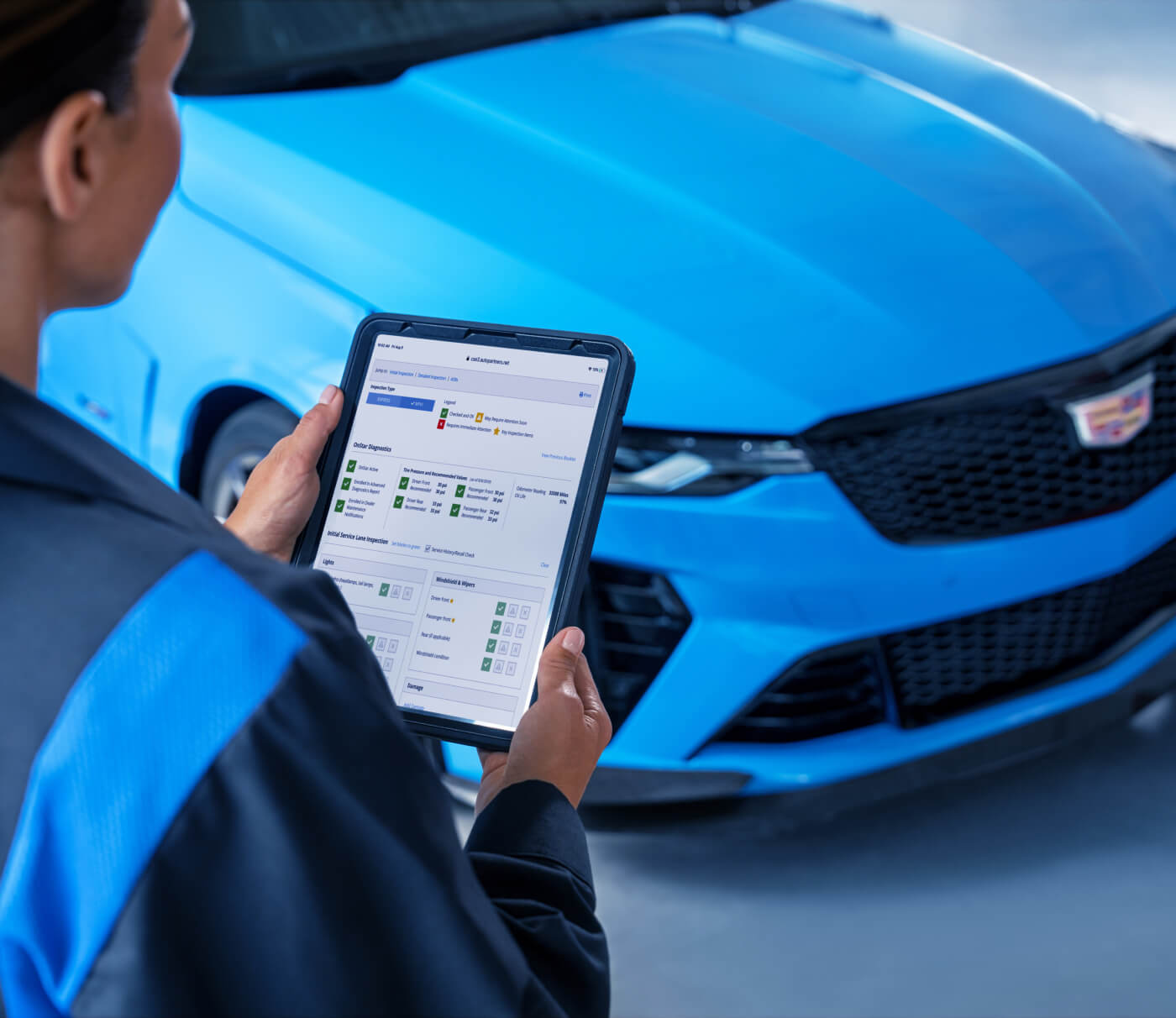 Front view of a technician holding a diagnostic tablet before a blue Cadillac in a service bay.
