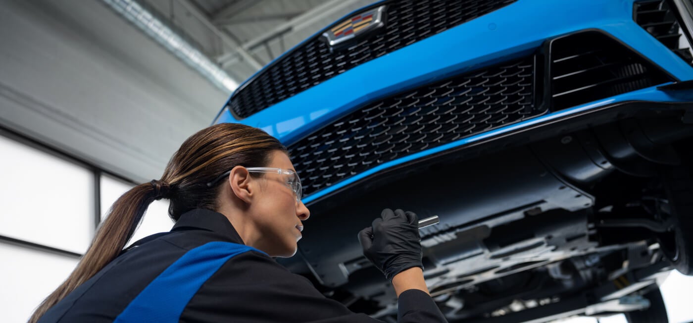Low-angle view of a technician inspecting the undercarriage of a lifted blue Cadillac with a flashlight.