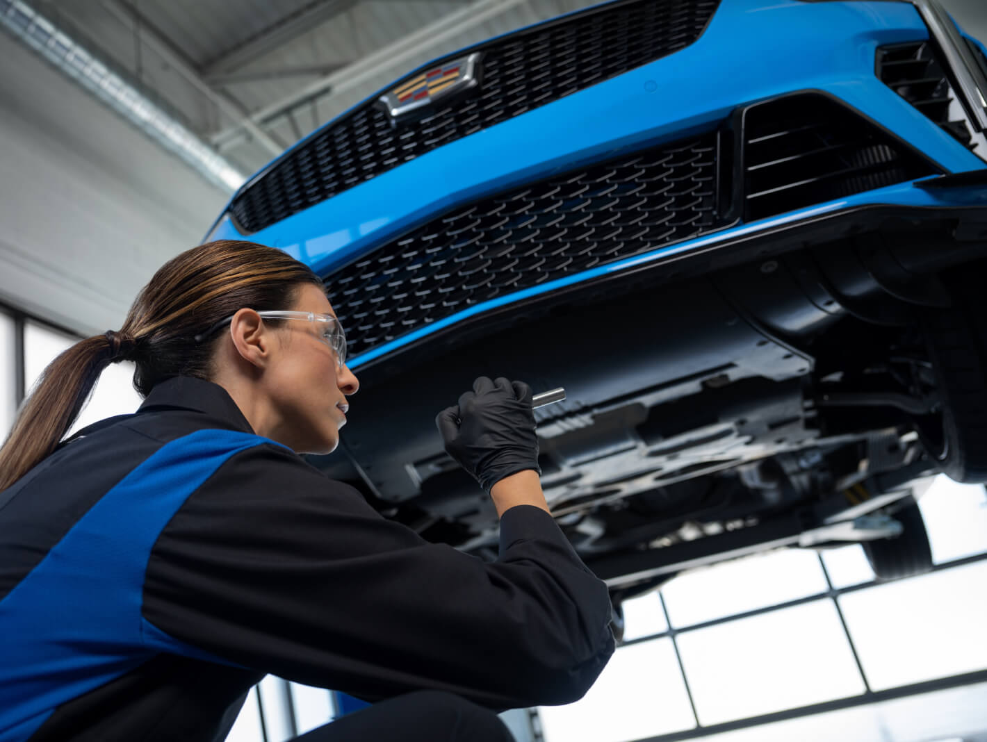 Low-angle view of a technician inspecting the underside of a lifted blue Cadillac CT4-V with a flashlight.