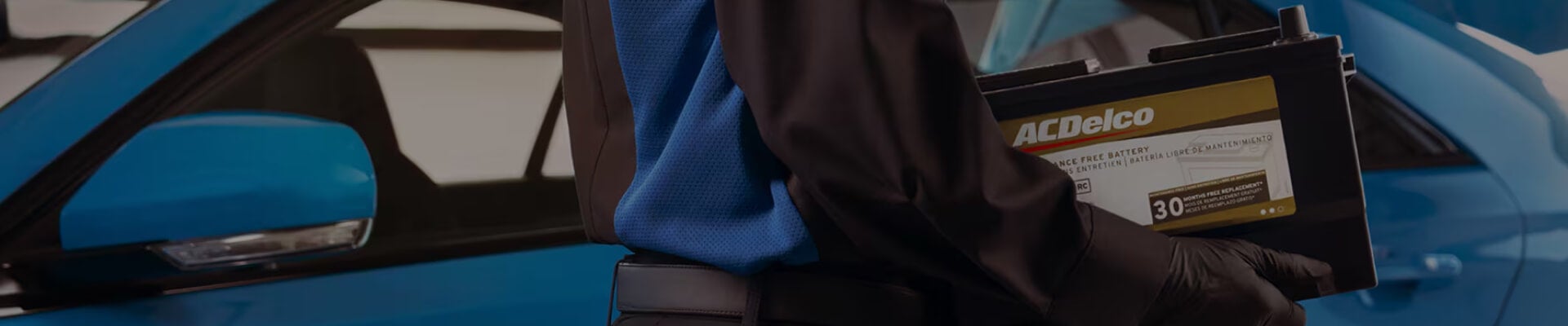 Side profile view of a female technician wearing safety glasses carrying an ACDelco battery next to a blue car with an open hood.