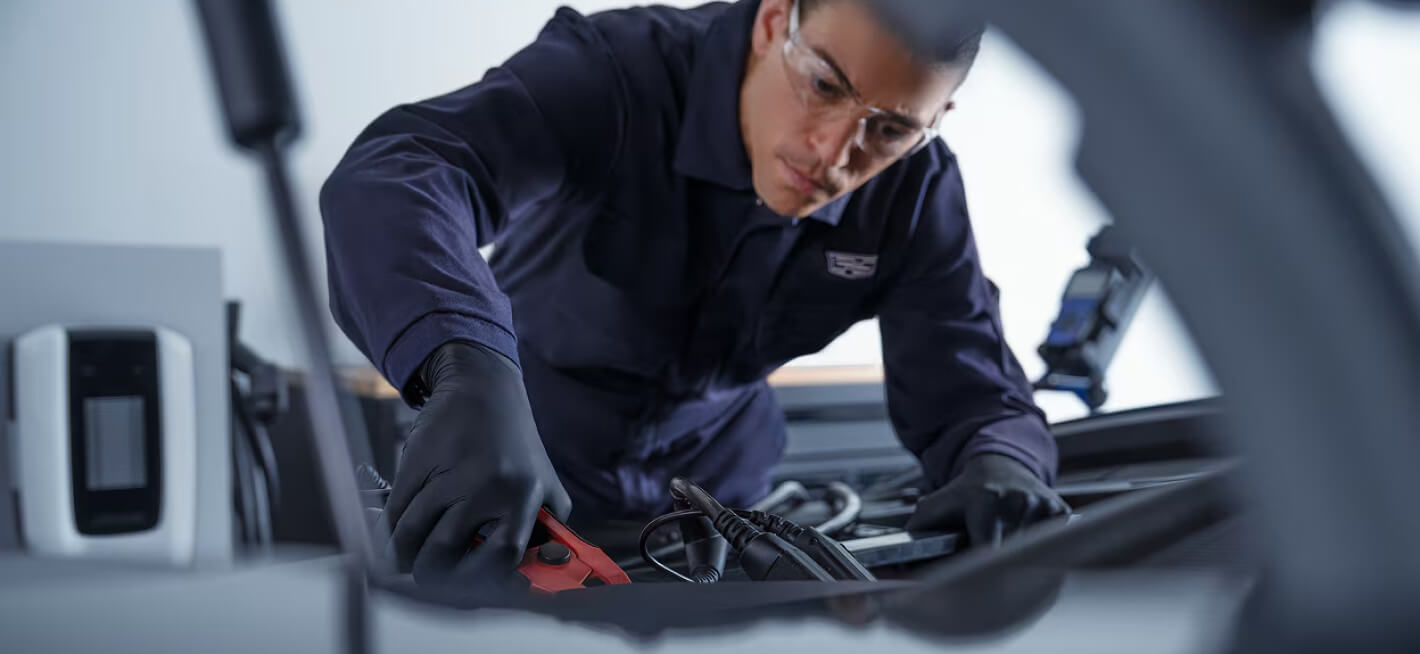 Close-up view of a male technician wearing safety glasses attaching jumper cables to a vehicle battery in a service bay.