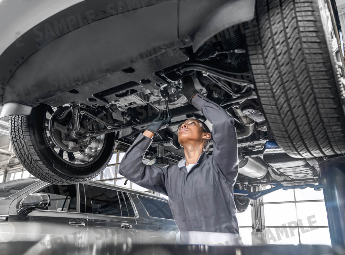 Low-angle view of a female technician wearing safety glasses working under a vehicle raised on a lift in a service bay.