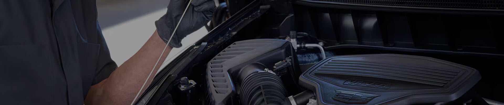 Side profile view of a male technician wearing safety glasses checking the oil dipstick under the hood of a vehicle engine bay.