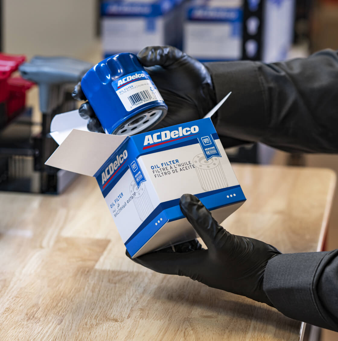 Close-up view of a technician wearing black gloves unboxing an ACDelco oil filter above a workbench.