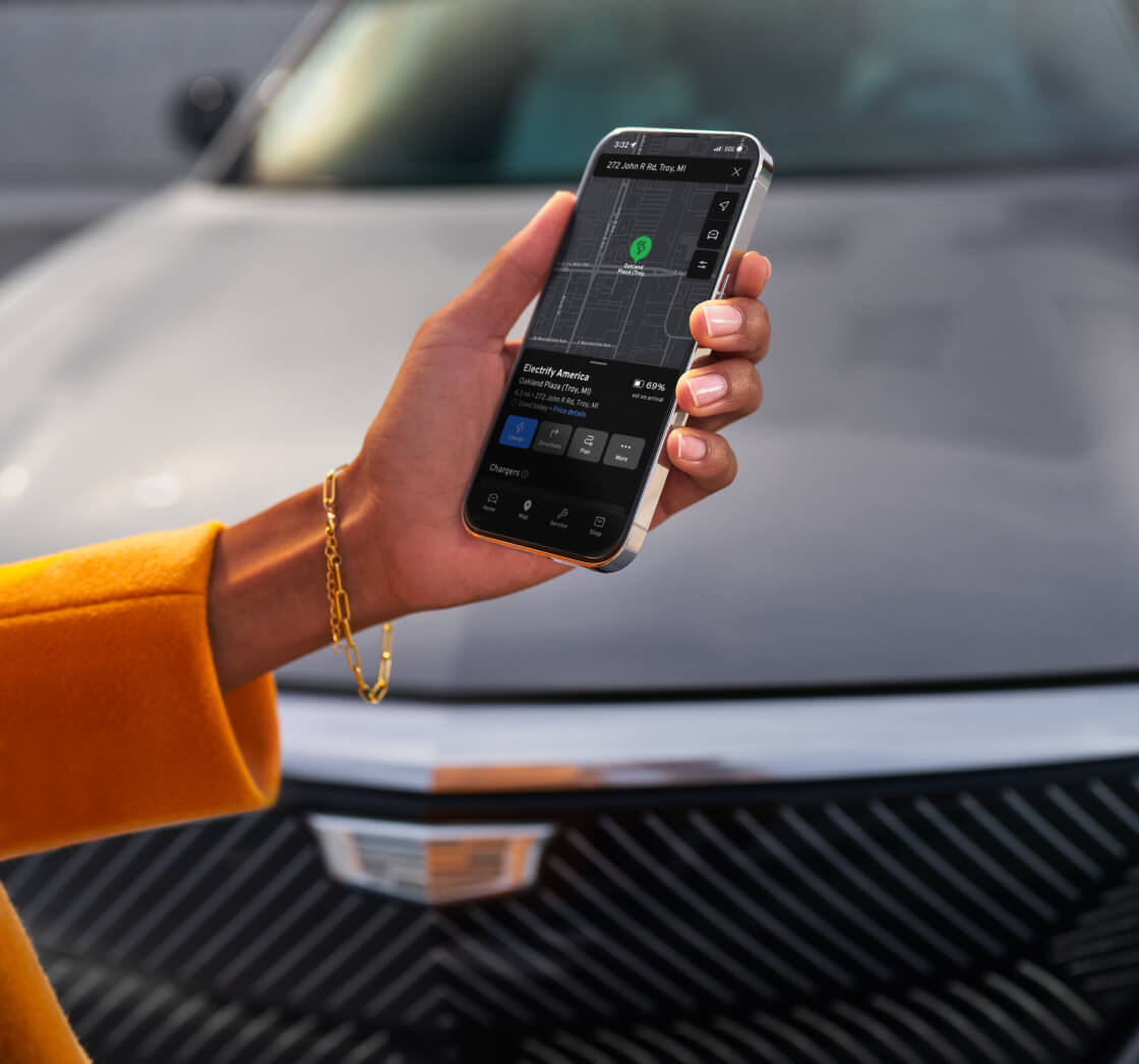 Close-up view of a woman wearing a yellow top holding a smartphone showing charging stations in front of a 2026 Cadillac LYRIQ.