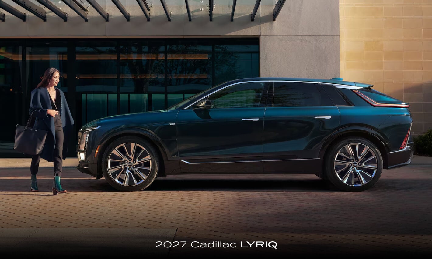 Exterior side view of a woman walking near a blue 2027 Cadillac LYRIQ parked on a sunny city street.